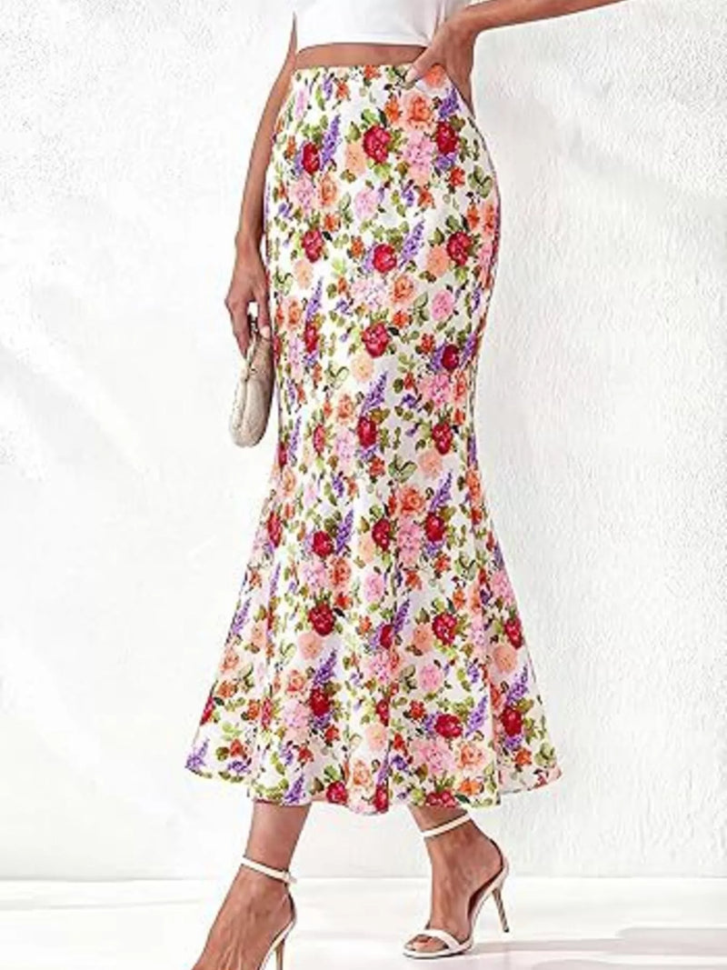 A woman models a high-waisted, flared midi skirt with a vibrant multicolor floral print on a white base. The skirt features pink, red, purple, and green flowers with green leaves.