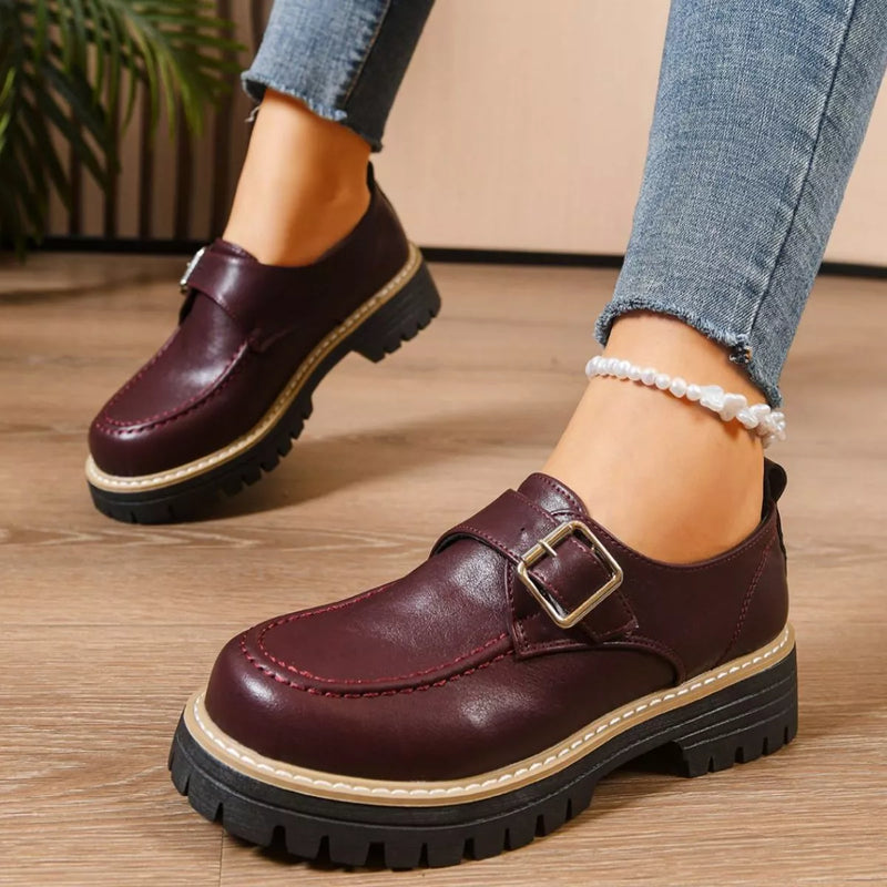 Brown leather shoes with buckles worn indoors on a wooden floor.