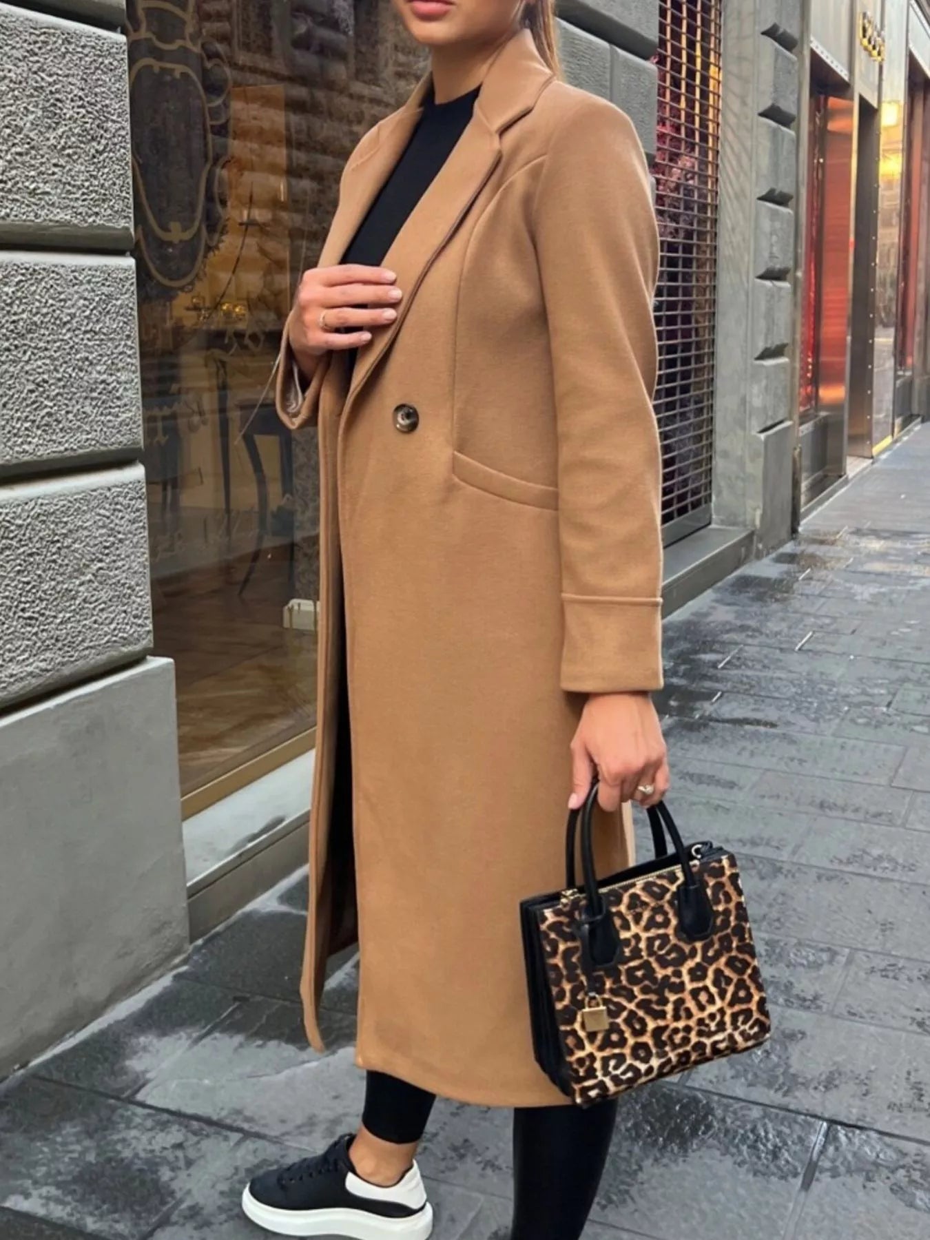 Person wearing a long beige coat holding a leopard print handbag on a city street.