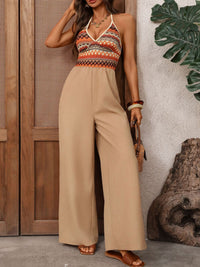 Woman wearing a patterned top and beige jumpsuit standing against a wooden wall with a plant.
