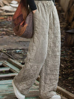 A person wearing loose, quilted wide-leg pants in a light taupe-beige color, standing outdoors on a weathered wooden platform. The pants have a diamond-quilted pattern and are paired with white sneakers and a brown patchwork shoulder bag.