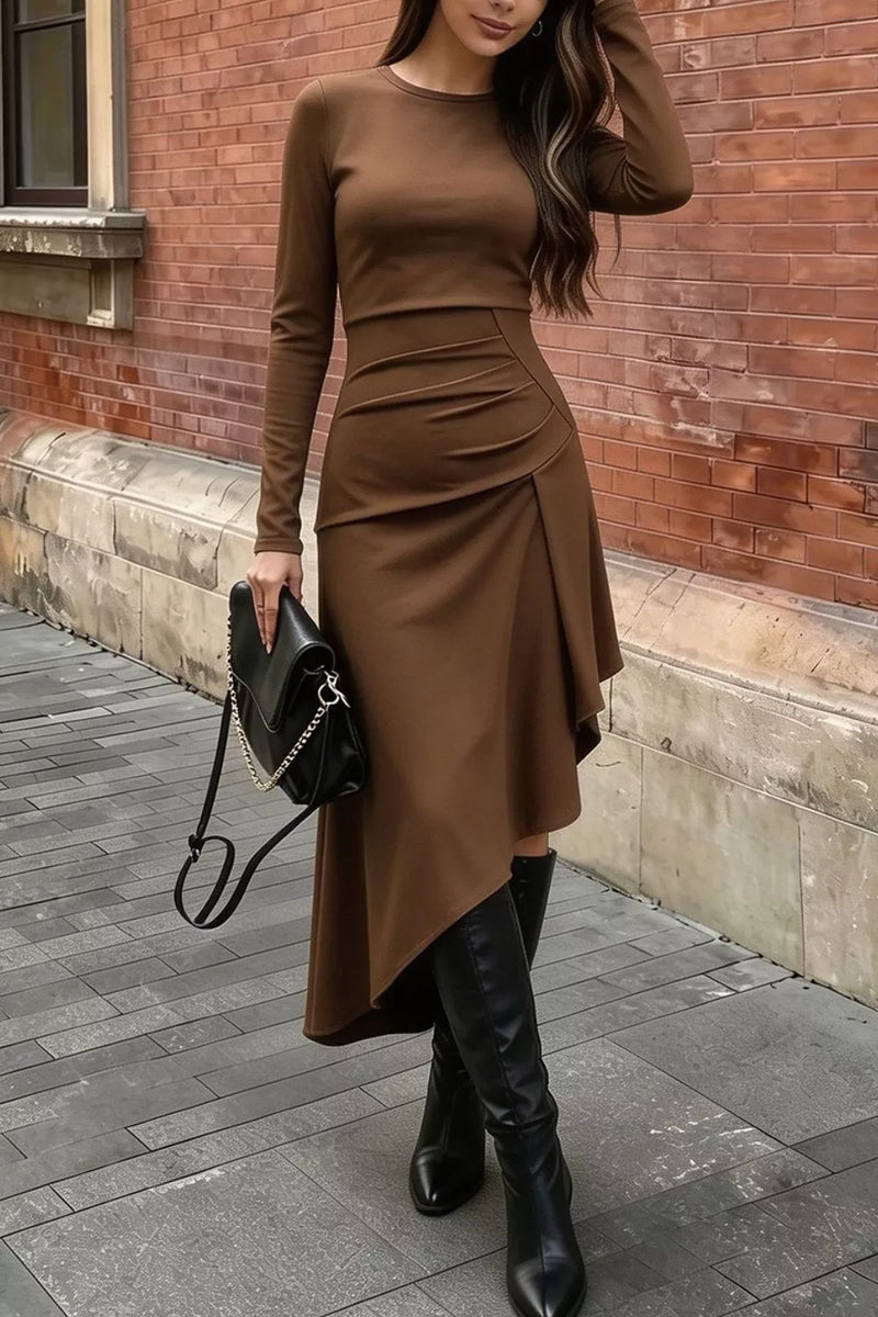 A woman wearing a fitted long-sleeve brown midi dress with an asymmetrical draped hem, styled with black knee-high boots and a black handbag, standing on a paved sidewalk in front of a red brick wall.
