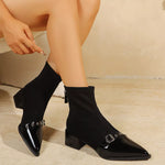A person seated indoors wearing black ankle boots with pointed toes, glossy patent leather detailing, and silver buckle hardware. The photo is cropped to the lower torso, legs, and feet against a warm beige wall and floor.