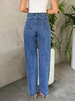 Person wearing blue jeans standing against a neutral wall with a plant in the background