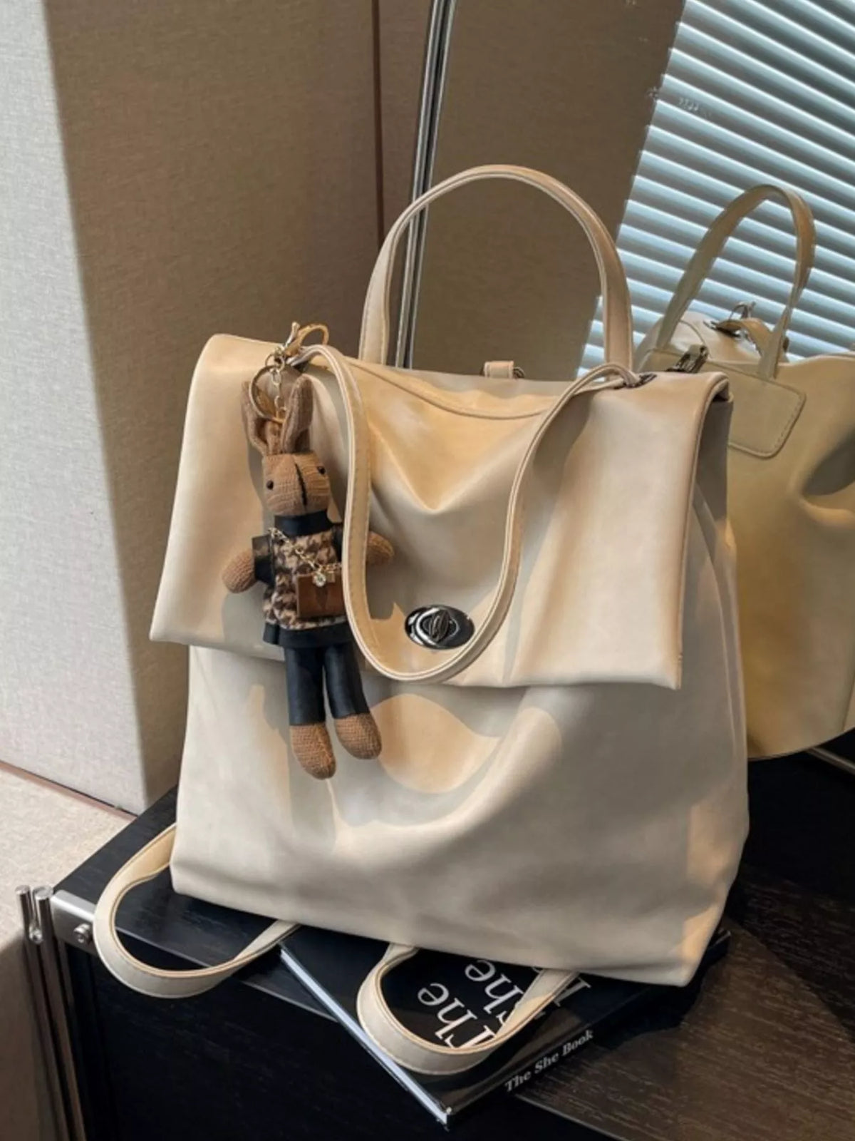 Beige handbag with a small plush toy attached, placed on a surface.