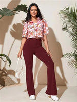 Woman wearing a floral top and burgundy pants with a white handbag, standing against a beige wall with plants.