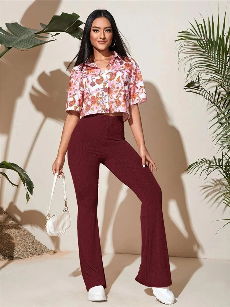 Woman wearing a floral top and burgundy pants with a white handbag, standing against a beige wall with plants.