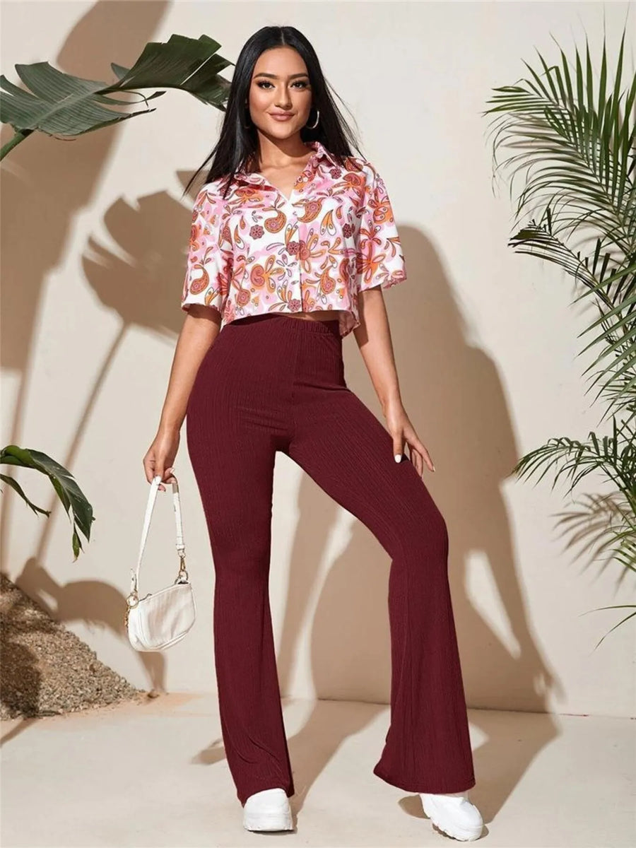 Woman wearing a floral top and burgundy pants with a white handbag, standing against a beige wall with plants.