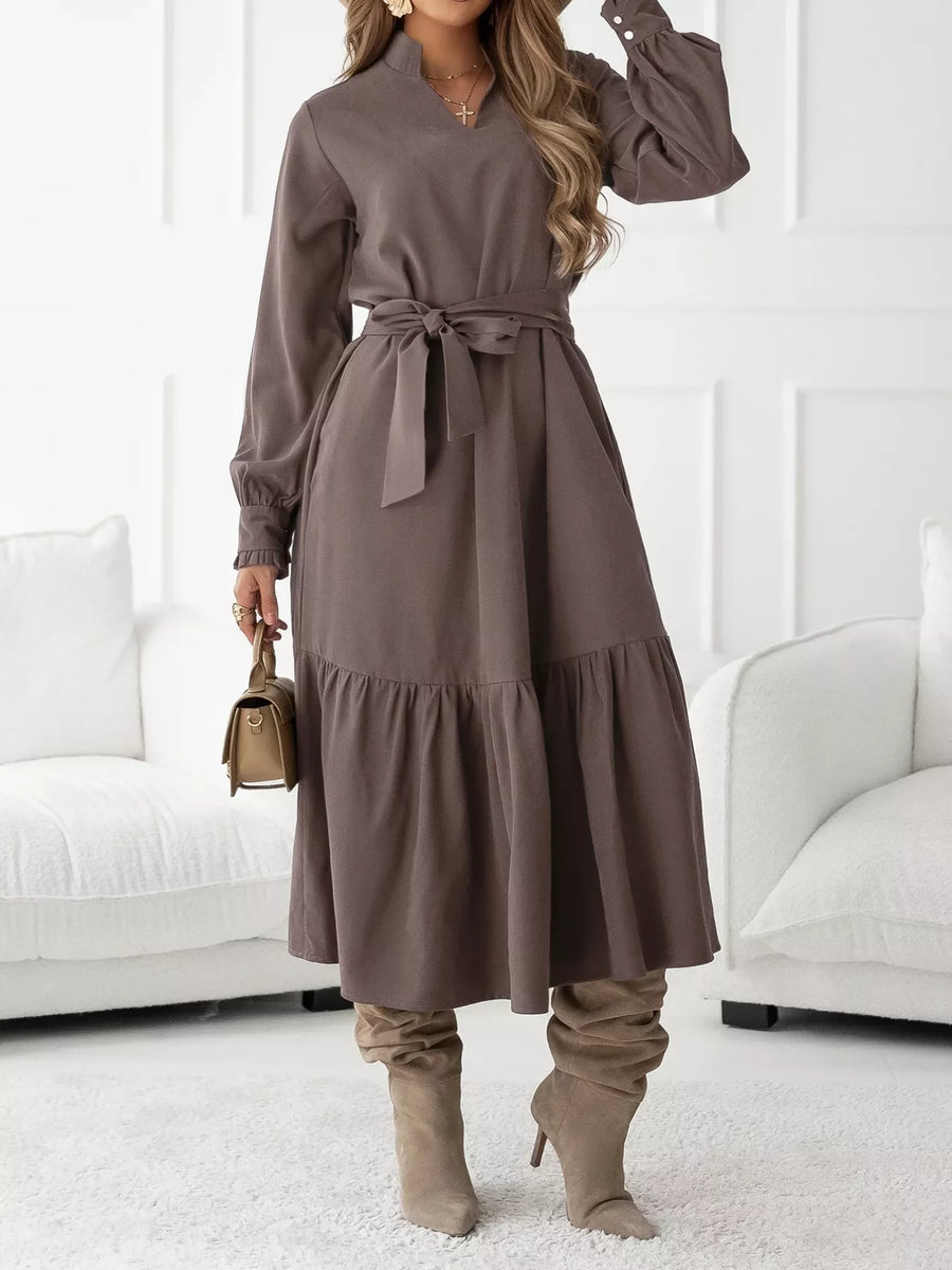 Woman wearing a brown dress with long sleeves and a belt, standing in a room with white furniture.