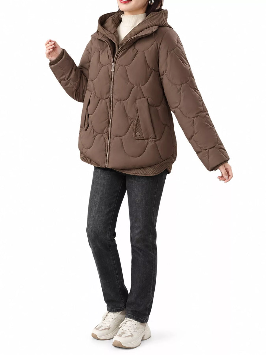 A woman wearing a brown quilted puffer jacket with a hood, paired with dark gray jeans and off-white sneakers, shown against a plain white background.
