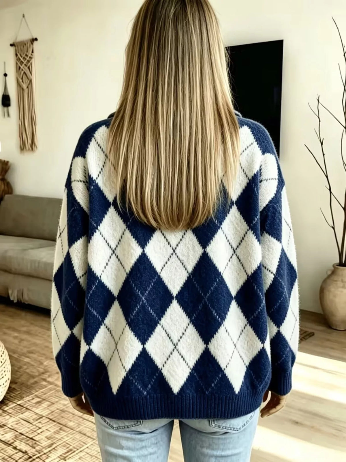 A woman viewed from behind wearing a navy blue and white argyle knit sweater with light blue jeans in a bright living room.