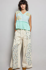 Woman wearing a light green top and cream pants with floral patterns against a gray background