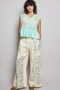 Woman wearing a light green top and cream pants with floral patterns against a gray background