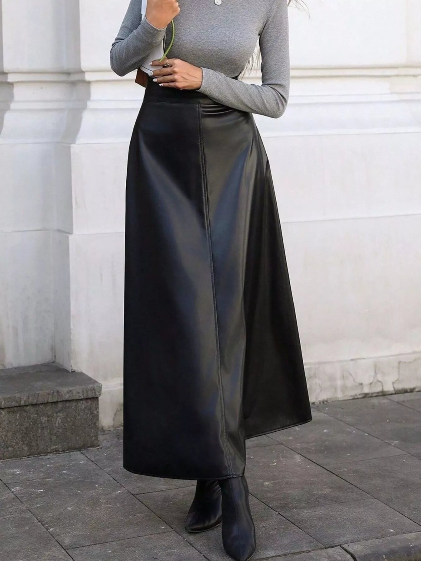 Person wearing a gray long-sleeve top and black leather skirt standing against a white wall.