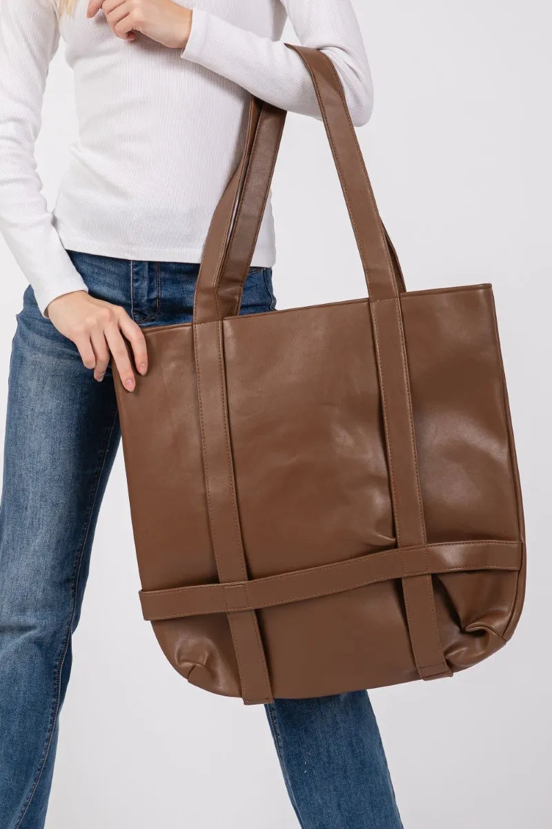 Brown leather tote bag held by a person wearing a white sweater and blue jeans on a light gray background