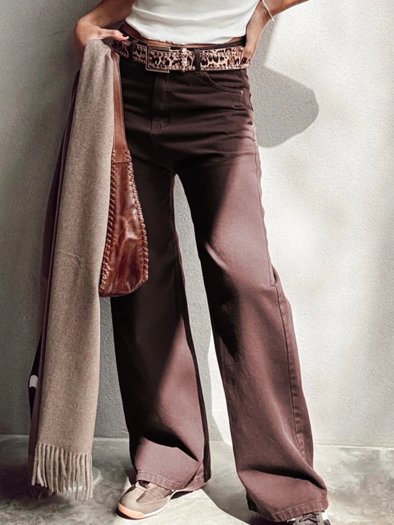 Person wearing wide-leg brown pants with a white shirt and patterned belt, holding a beige scarf.