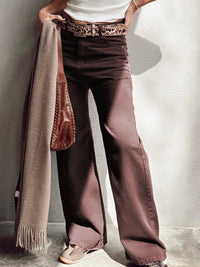 Person wearing wide-leg brown pants with a white shirt and patterned belt, holding a beige scarf.