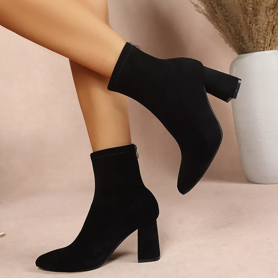 Black suede ankle boots with block heels, shown on a model's feet against a soft beige background with a white vase of dried grass.
