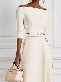 Woman wearing a white off-shoulder dress with a belt, holding a beige handbag against a white background.