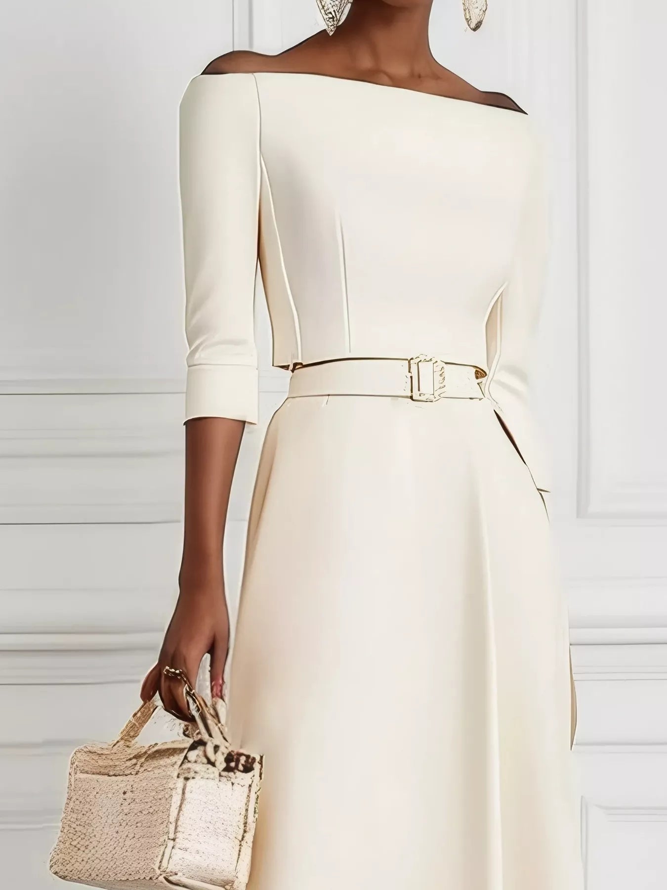 Woman wearing a white off-shoulder dress with a belt, holding a beige handbag against a white background.