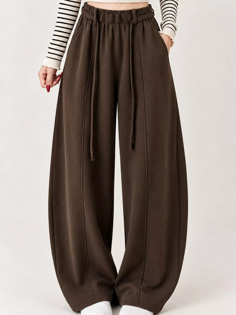 A person wearing loose, wide-leg brown pants with a drawstring waistband, paired with a black-and-white striped long-sleeve top, shown against a plain light background.