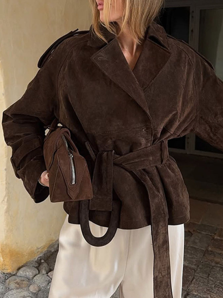 Person wearing a brown suede coat with a matching handbag, standing against a neutral wall.
