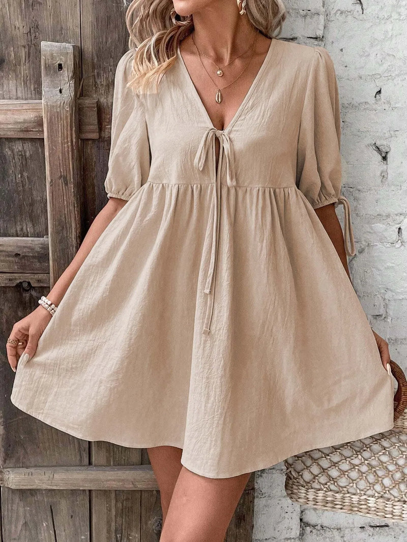 Woman wearing a beige dress standing against a rustic wooden and brick wall.