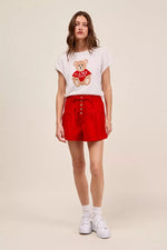 Woman wearing a white t-shirt with a teddy bear graphic and red shorts on a beige background