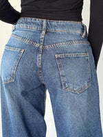 Close-up back view of a person wearing blue denim jeans with a black long-sleeve top, showing the back pockets and waistband details.