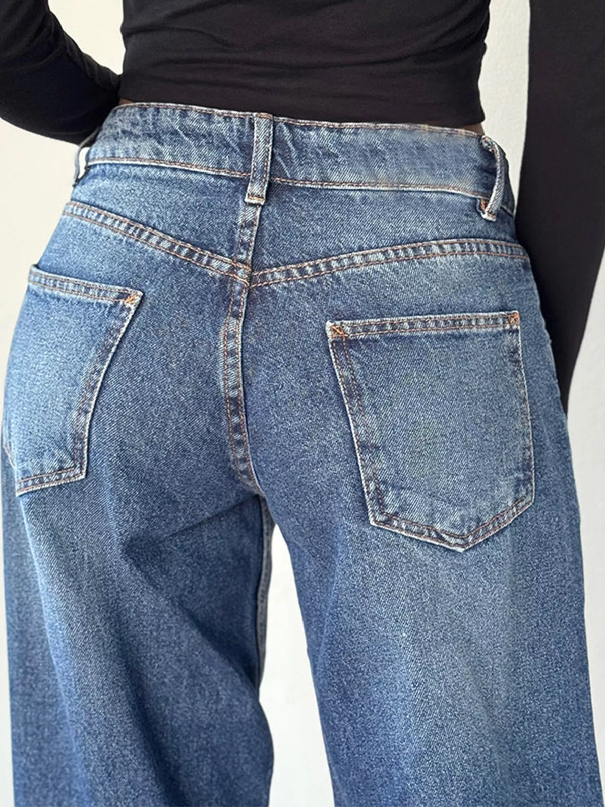 Close-up back view of a person wearing blue denim jeans with a black long-sleeve top, showing the back pockets and waistband details.