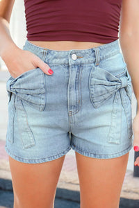 Person wearing light blue denim shorts with a bow detail, standing against a blurred background.