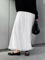 Person wearing a black top, white skirt, and black shoes against a gray wall.