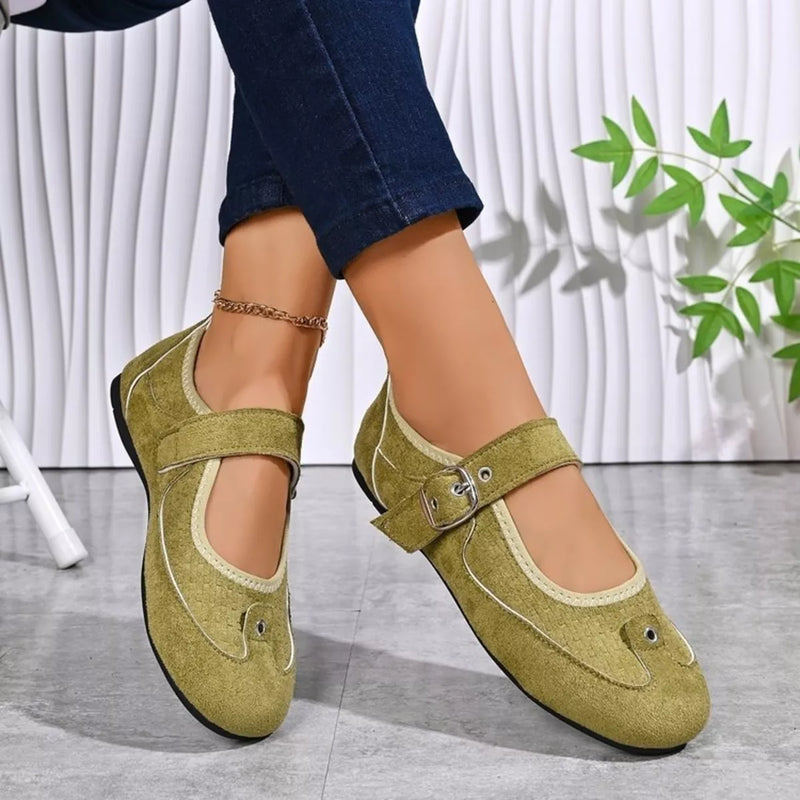A pair of women's Mary Jane shoes in olive green suede, shown being worn with dark blue jeans. The shoes feature a single strap across the instep with a silver buckle, a decorative stitched detail on the toe, and a low block heel, set against a modern white textured wall and gray floor.