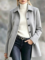 A woman wearing a light gray wool coat over a cream ribbed turtleneck sweater and dark blue jeans, with a black belt featuring a gold buckle.