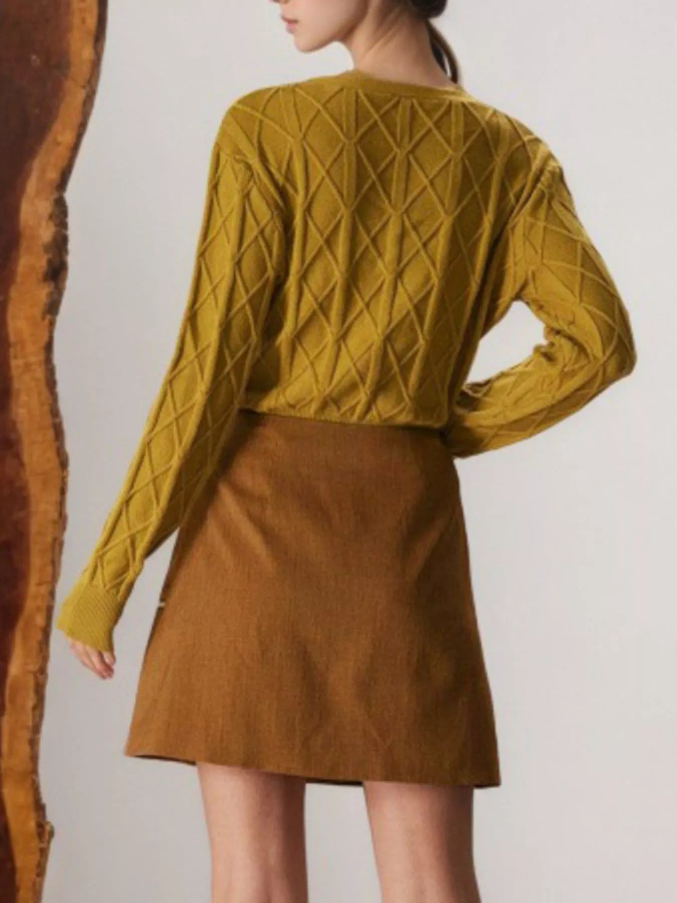 Person wearing a mustard yellow knitted sweater and brown skirt on a plain background