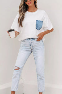 Person wearing a white t-shirt with a denim pocket design and light blue jeans, holding a coffee cup.