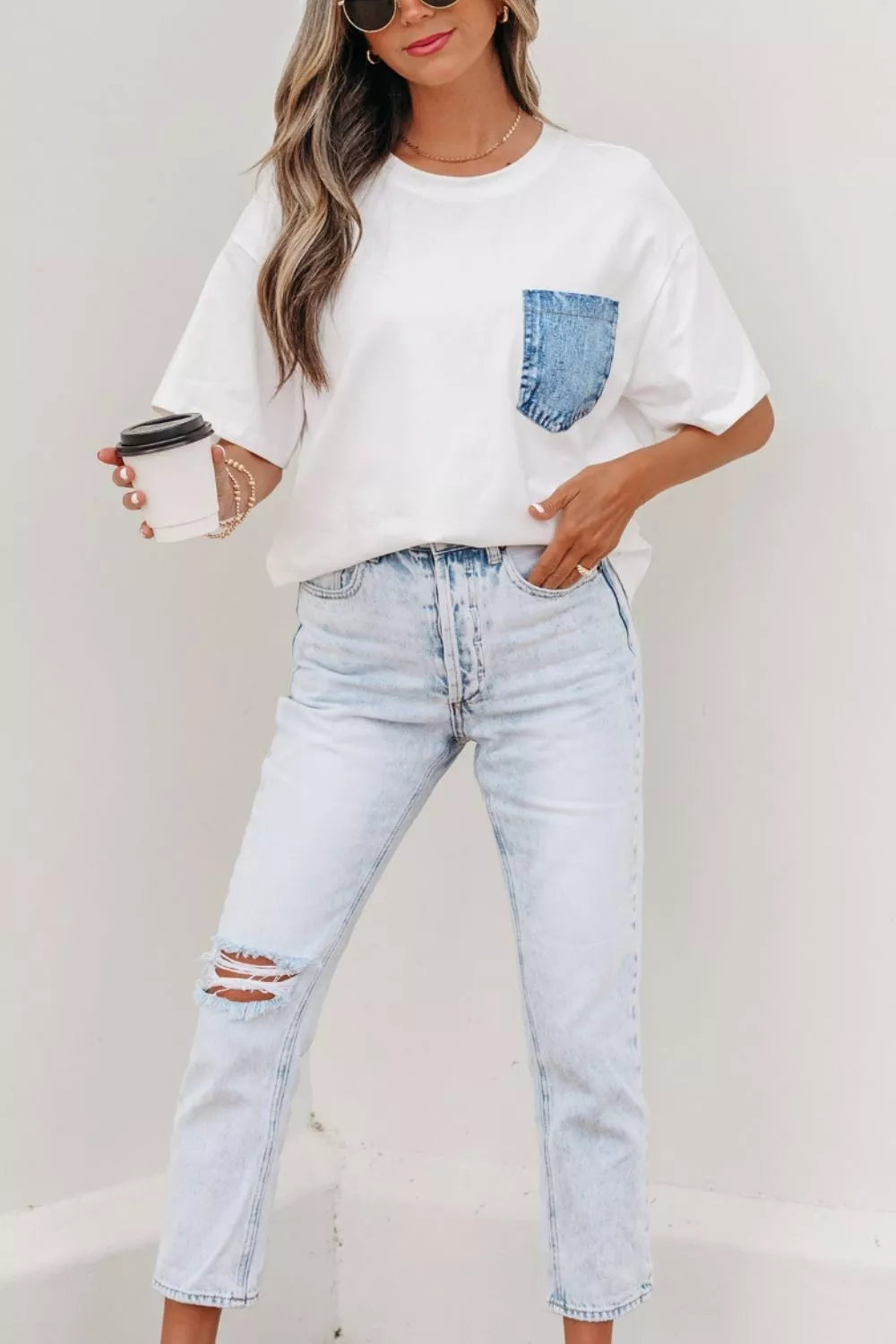 Person wearing a white t-shirt with a denim pocket design and light blue jeans, holding a coffee cup.