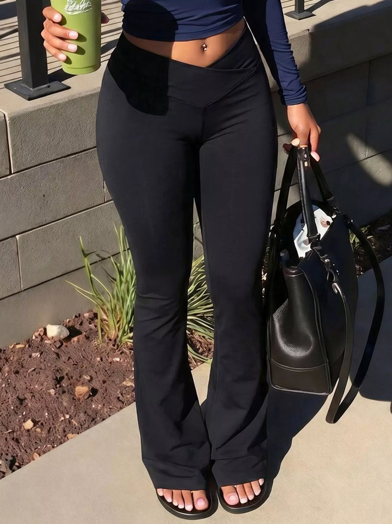 A person wearing black high-waisted flare leggings and a navy long-sleeve crop top, standing outdoors on a concrete walkway and holding a green coffee cup and a black tote bag.