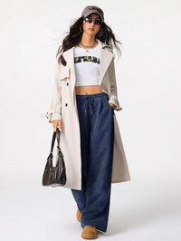 Woman wearing a long beige coat, white crop top, blue jeans, and brown boots on a white background