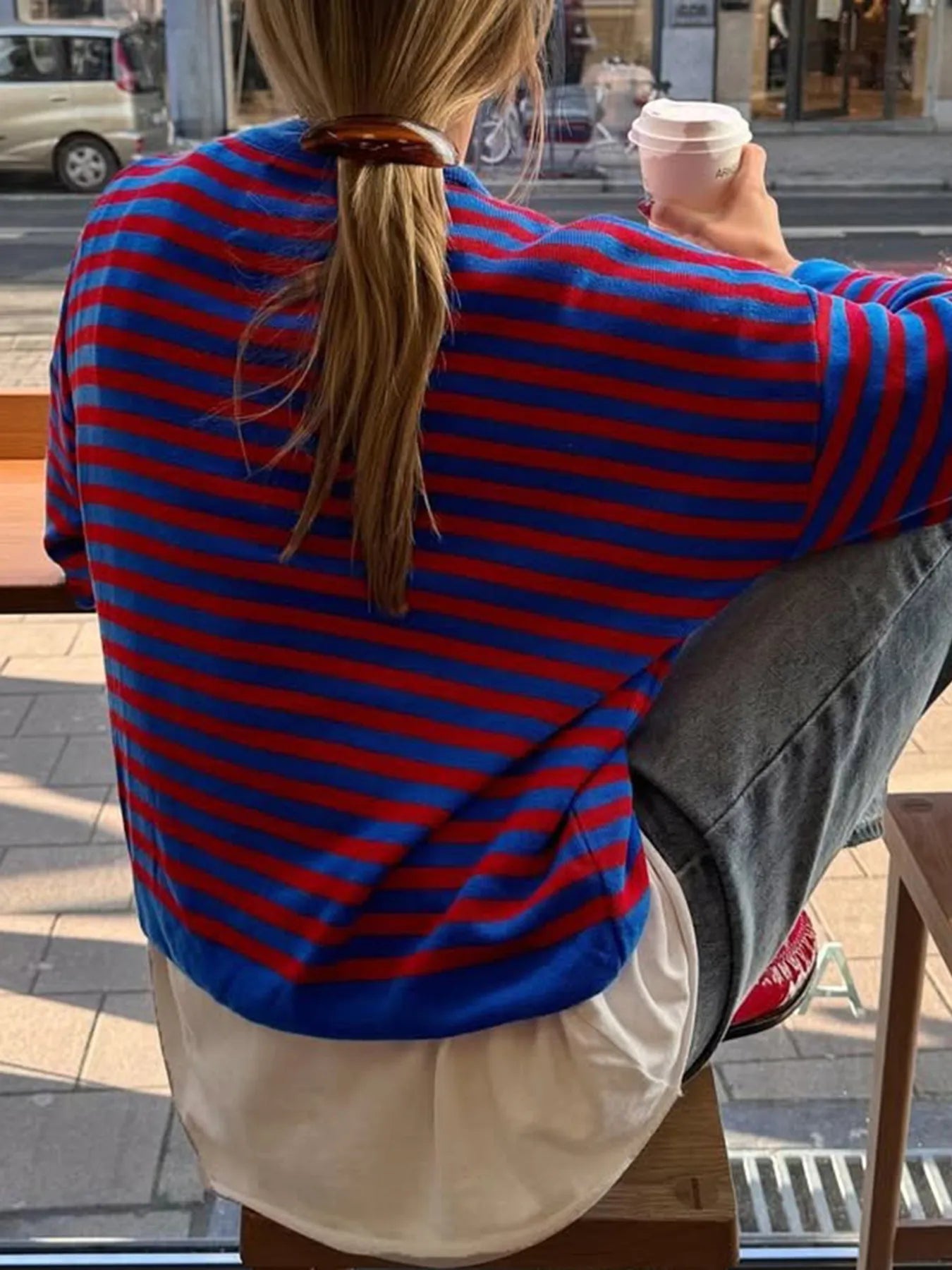 Person wearing a blue and red striped shirt sitting outdoors holding a cup.