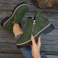 Green suede ankle boots with zippers held by a hand on a wooden floor.