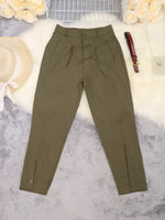 Olive green pleated trousers laid flat on a white faux fur surface with decorative props including a straw hat, flowers, a small bag, and a brown belt.