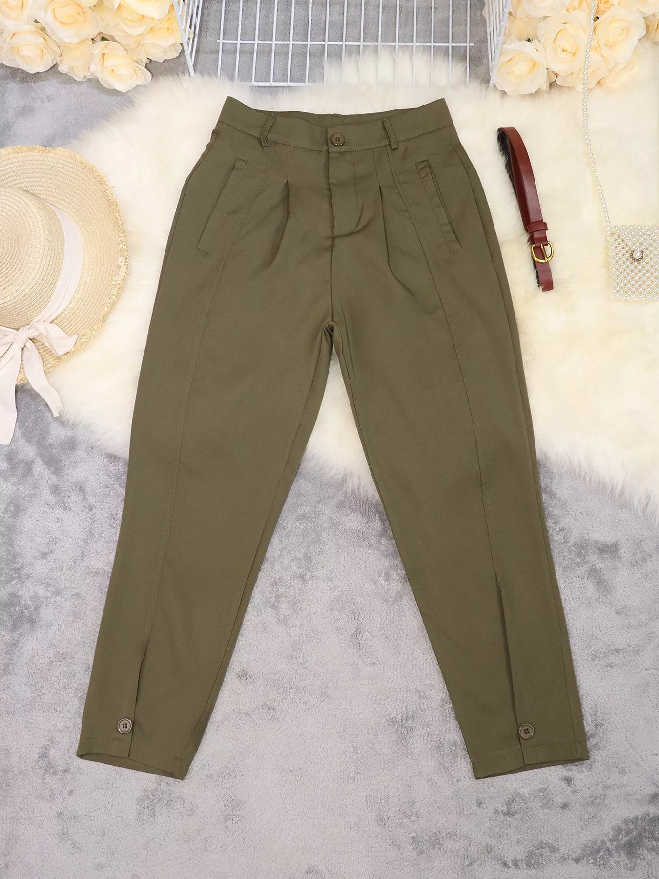 Olive green pleated trousers laid flat on a white faux fur surface with decorative props including a straw hat, flowers, a small bag, and a brown belt.