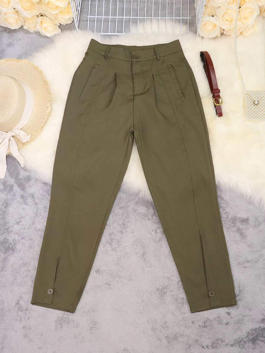 Olive green pleated trousers laid flat on a white faux fur surface with decorative props including a straw hat, flowers, a small bag, and a brown belt.