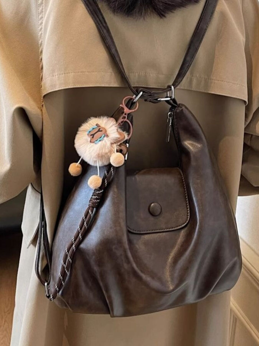 Brown leather handbag with decorative elements worn by a person in a beige coat.