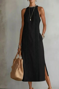 Woman wearing a black sleeveless dress holding a beige handbag against a plain background