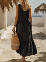 Woman in a black dress holding a straw bag on a beach with white buildings and blue sea in the background.