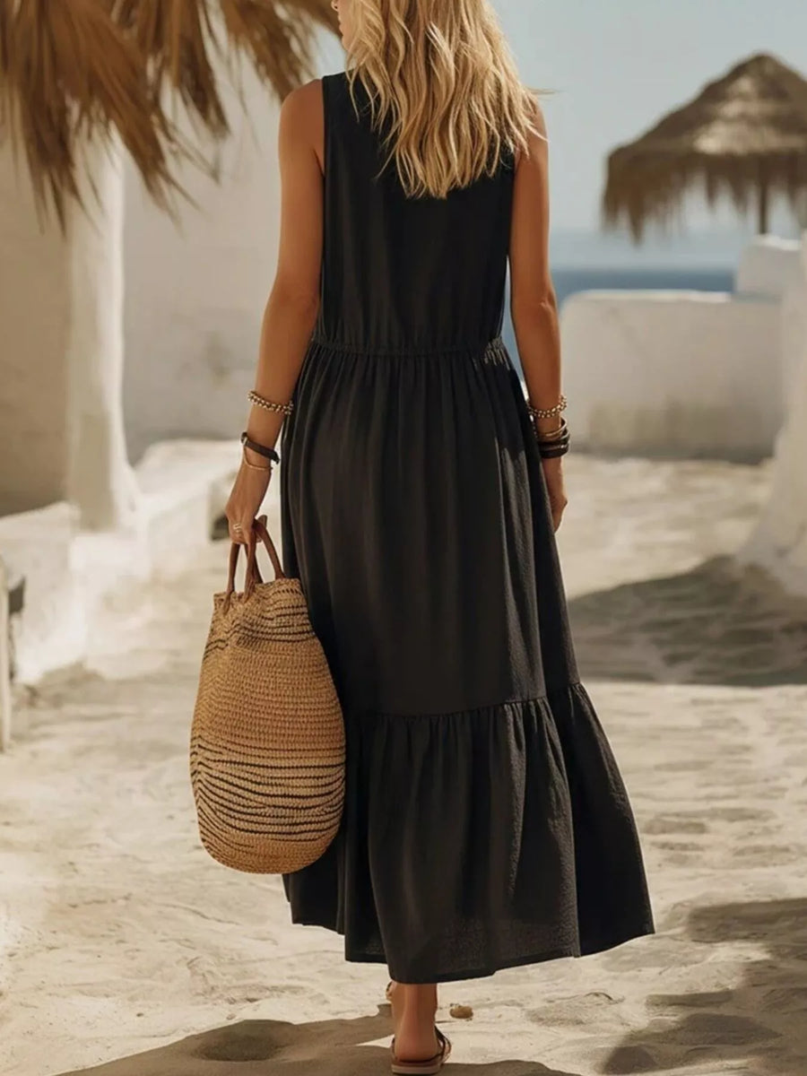 Woman in a black dress holding a straw bag on a beach with white buildings and blue sea in the background.