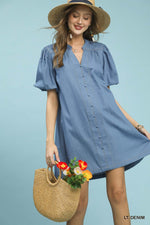 Woman wearing a light blue dress with a straw hat and holding a woven bag with flowers against a light blue background