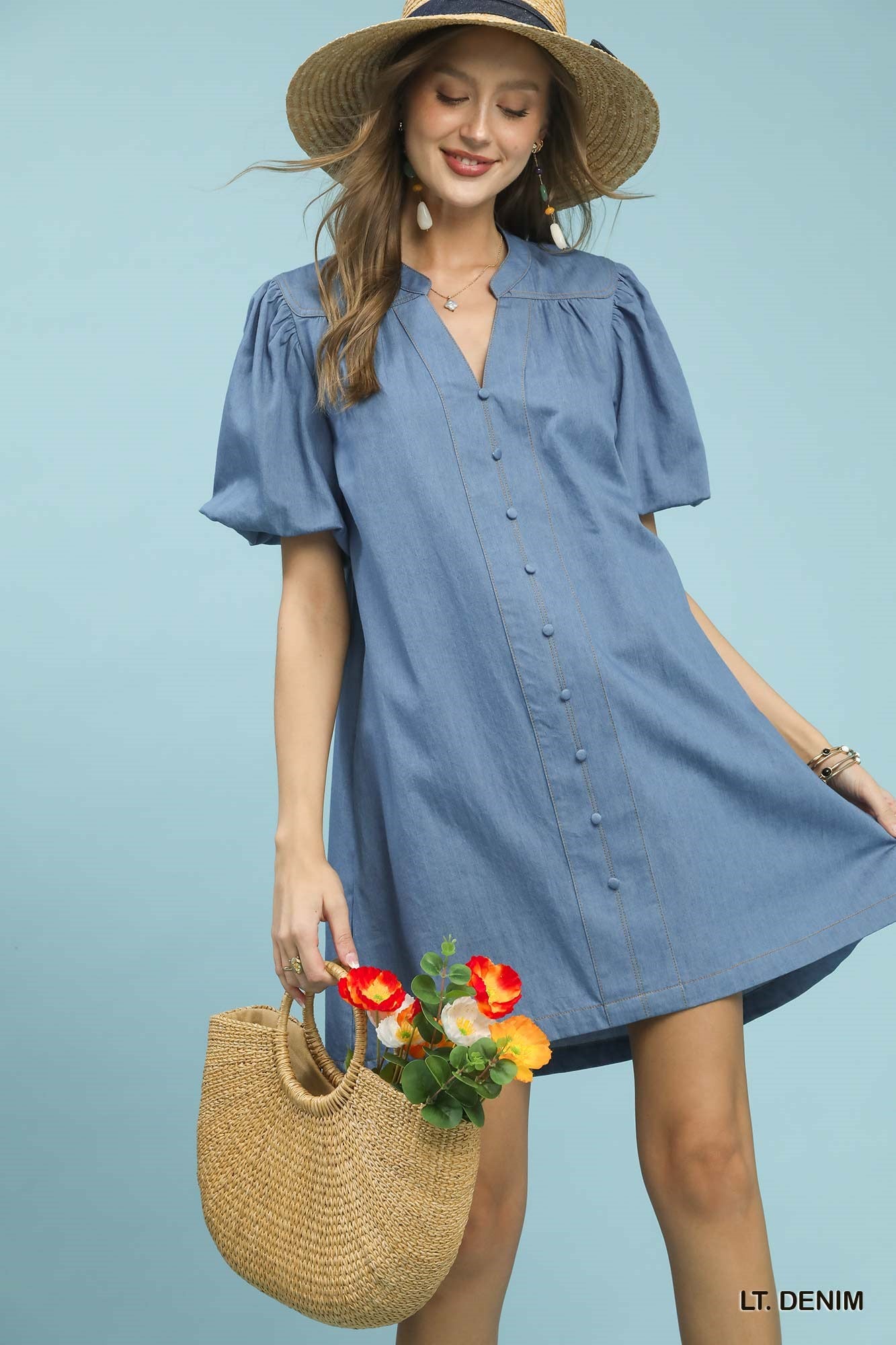 Woman wearing a light blue dress with a straw hat and holding a woven bag with flowers against a light blue background